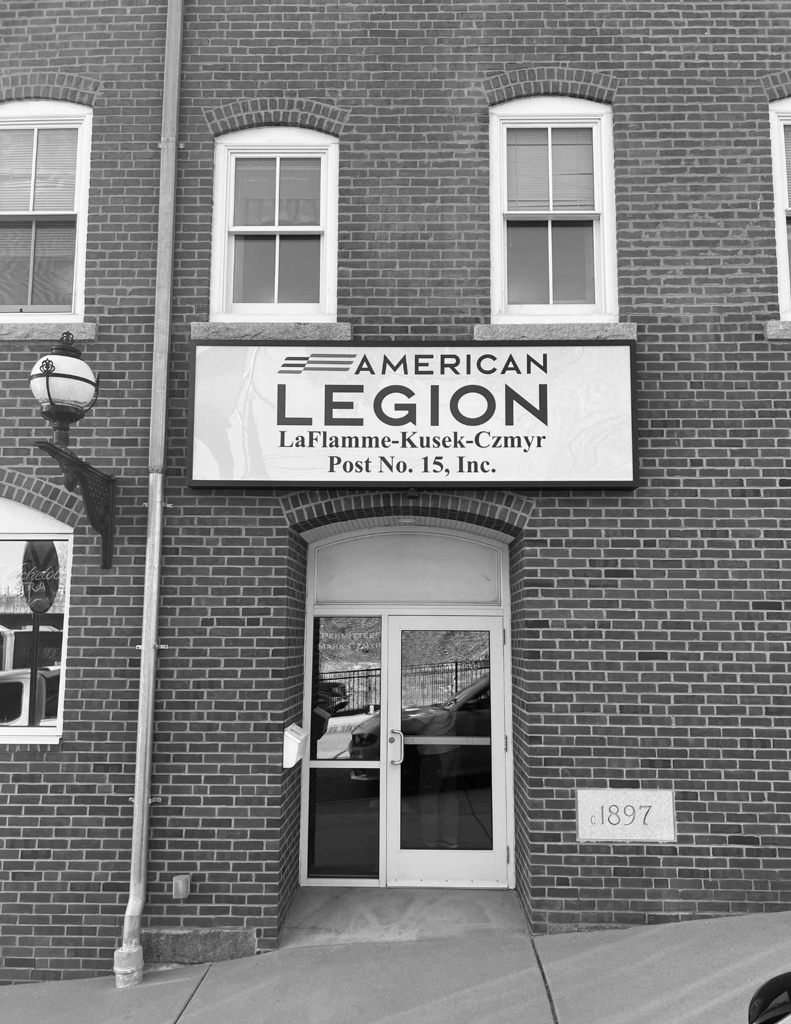 Brick building with a sign reading 'American Legion LaFlamme-Kusek-Czmur Post No. 15, Inc.' above the entrance. The entrance has a glass door. There are windows above the sign and a lamp post next to the building.