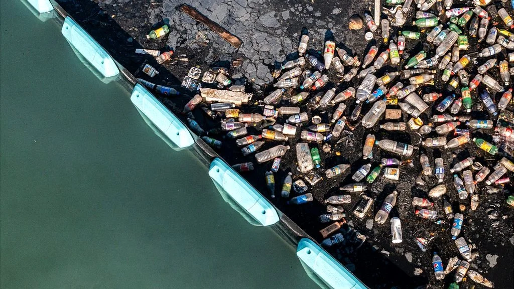 Hundreds of discarded plastic bottles lie on blackened ground near a body of water, with a barrier separating the two areas.