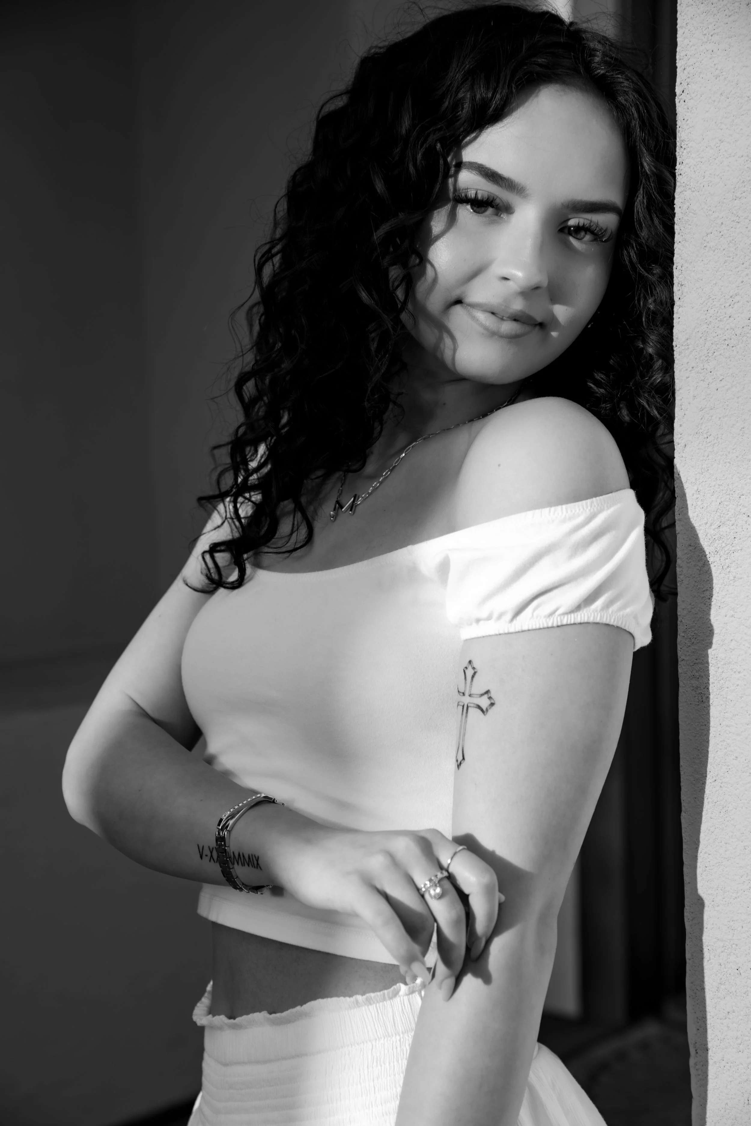 A young woman with dark, curly hair smiling at the camera, leaning against a wall. She is wearing an off-shoulder top, jewelry, and has visible tattoos on her arm. The photo is in black and white.