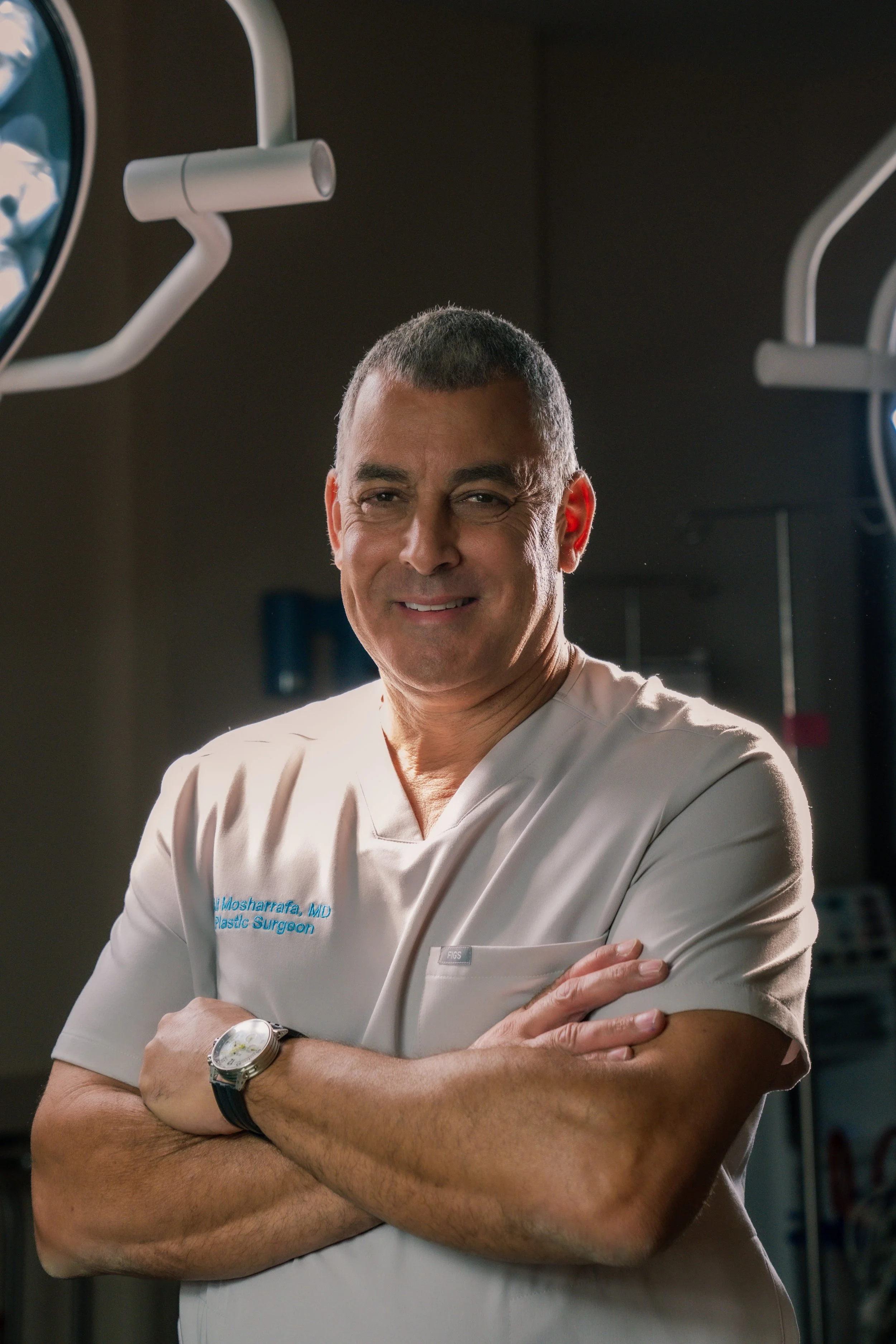 A male surgeon in scrubs standing in a hospital operating room with arms crossed and smiling at the camera.