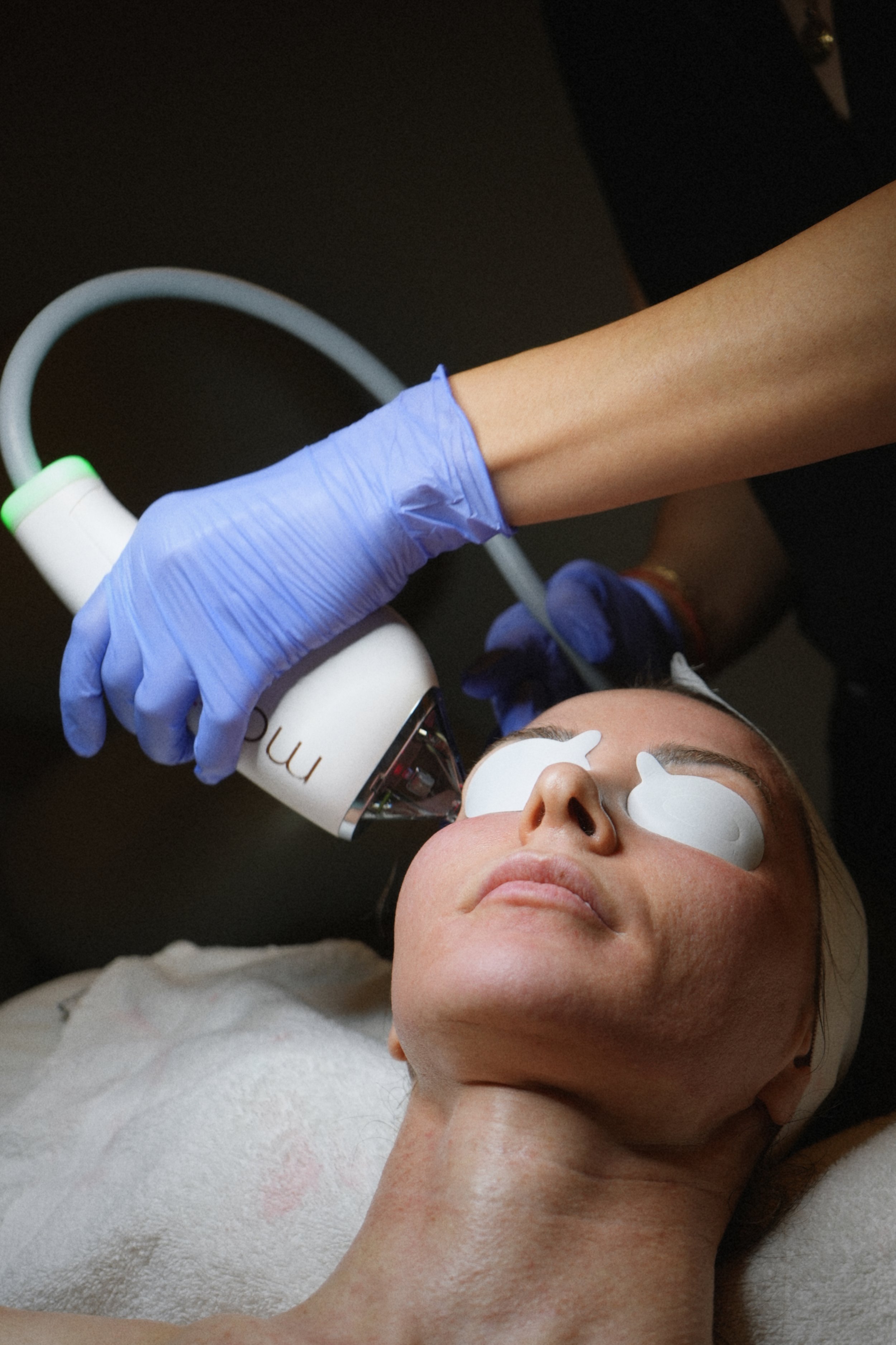 A woman lying down with eye patches, receiving a facial treatment from a professional using a handheld device.