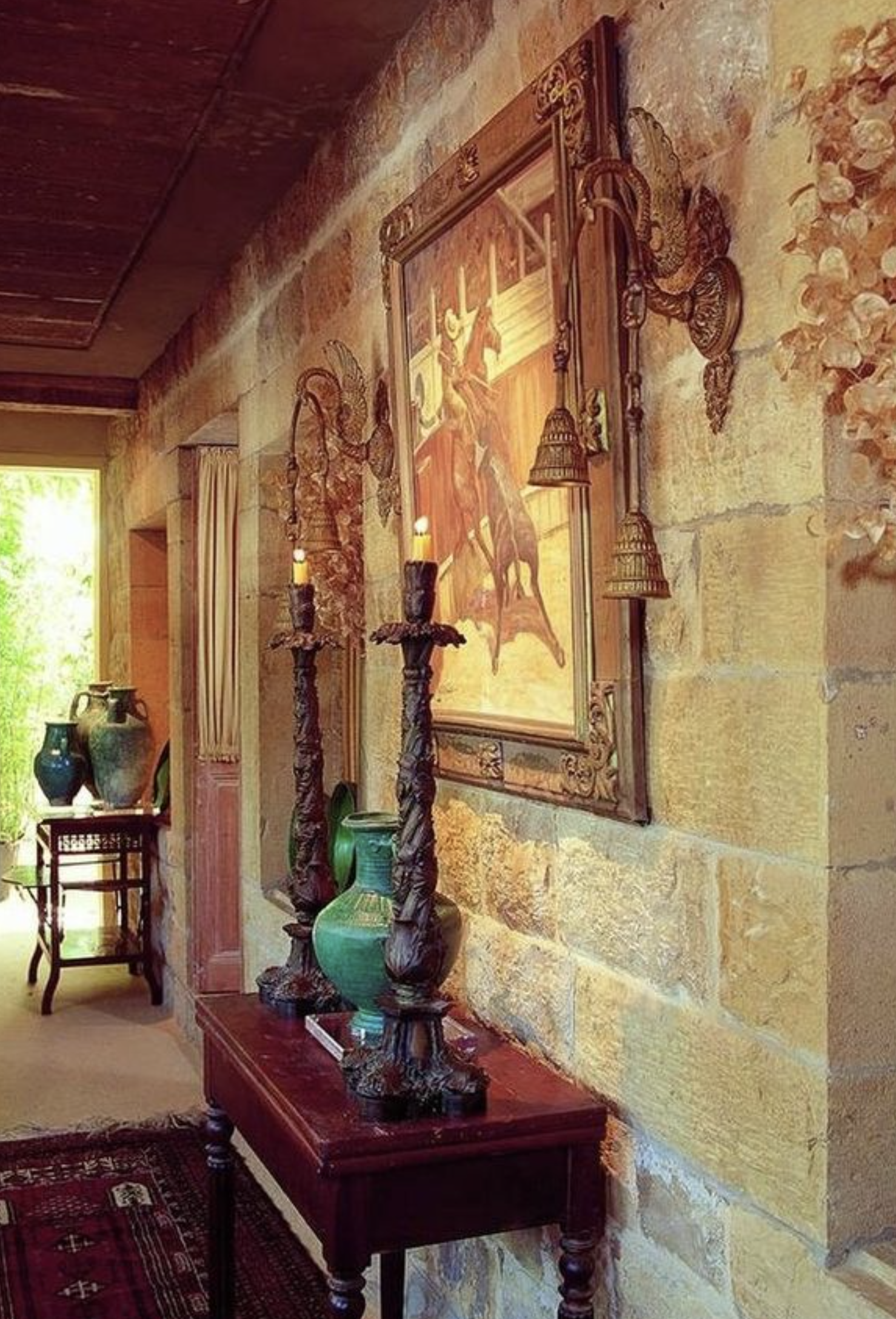 Decorative wooden table with two intricately carved candlesticks and a large green vase, against a stone wall with framed artwork, in a sunlit room.