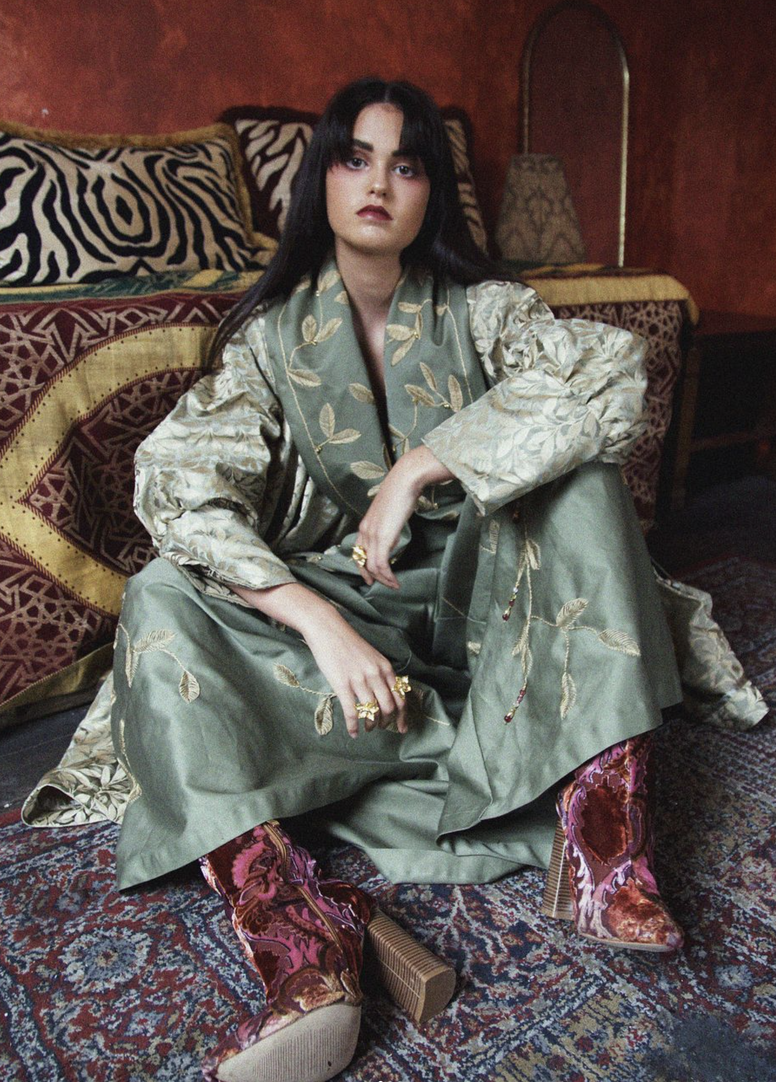 A woman with long dark hair, sitting on a patterned rug with a serious expression, wearing a patterned green and beige dress with embroidered leaves, and pink velvet velvet pants with floral motifs, in a room with a red wall, ornate pillows, and a floor lamp.