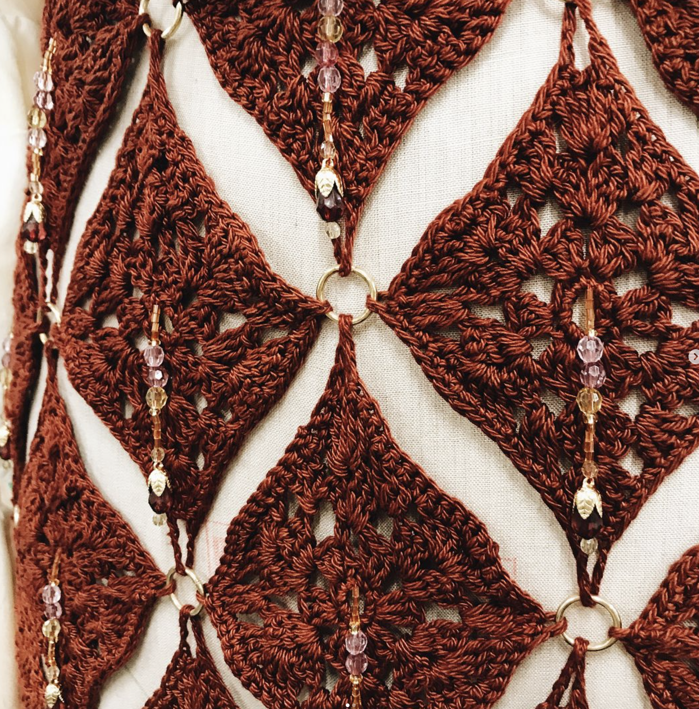 Close-up of a decorative textile with intricate red crochet patterns and hanging beads and rings on a light background.
