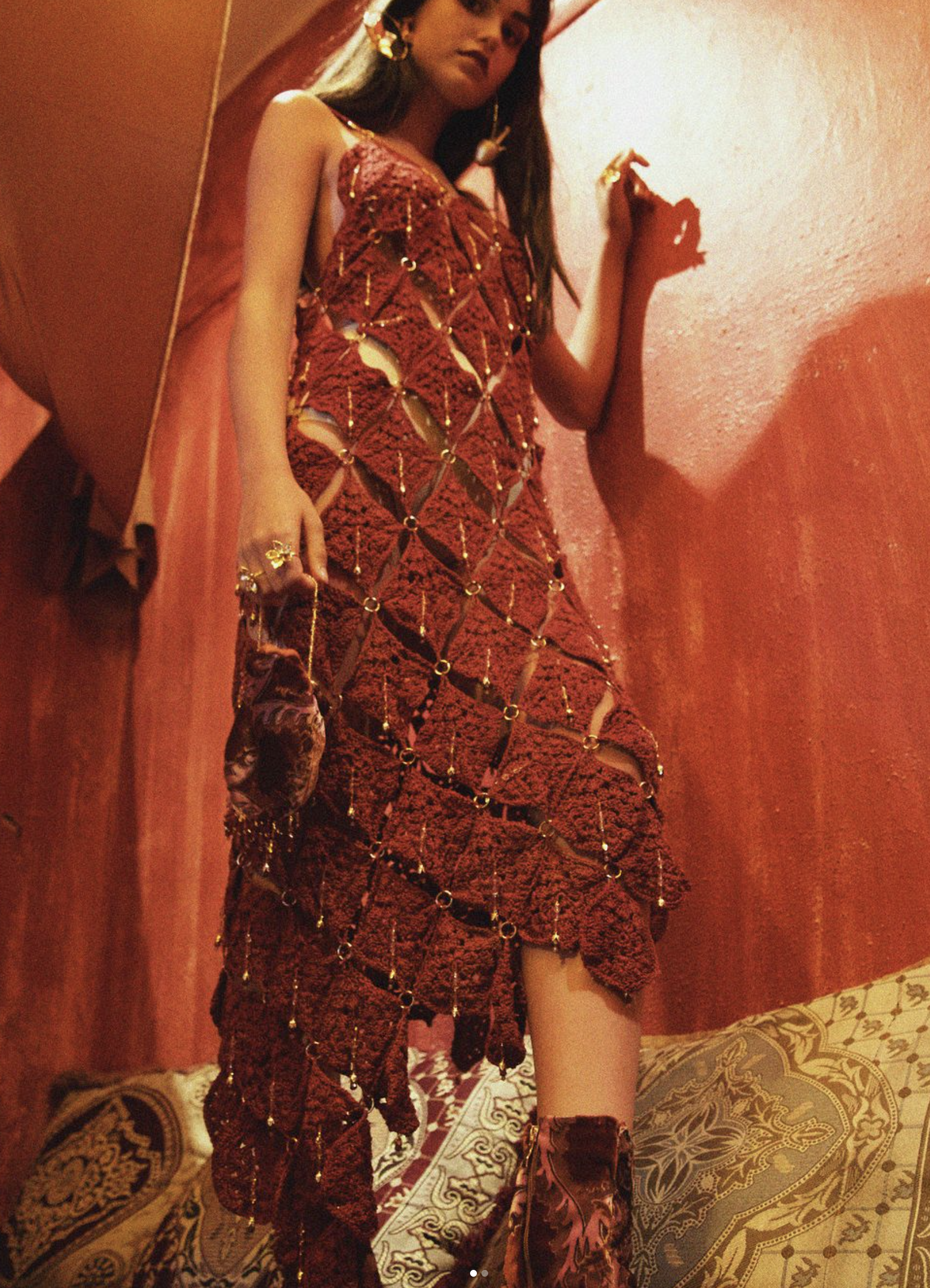 A woman dressed in a red, netted outfit with gold embellishments standing against a red wall. She is holding a small, velvet bag and wearing patterned shoes, jewelry, earrings, and a necklace. The setting appears to be indoors with decorative pillows and a patterned floor.