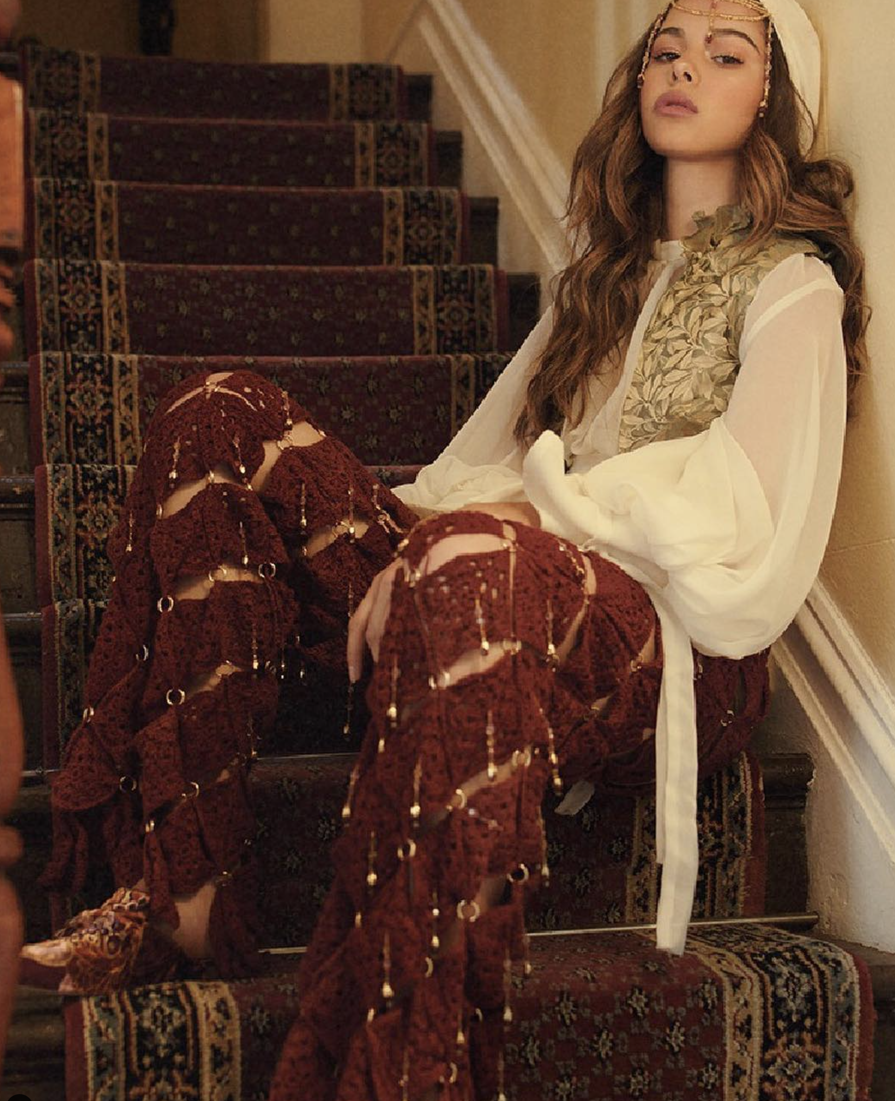 A woman with long hair, wearing a headscarf, sitting on a staircase with a patterned carpet. She is dressed in a white outfit and red crochet footwear.