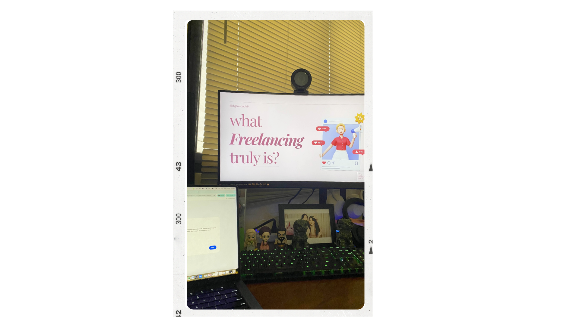 Computer desk setup with a monitor displaying the question 'what freelancing truly is?', a laptop with an open webpage, figurines, and a small framed photo, with window blinds in the background.