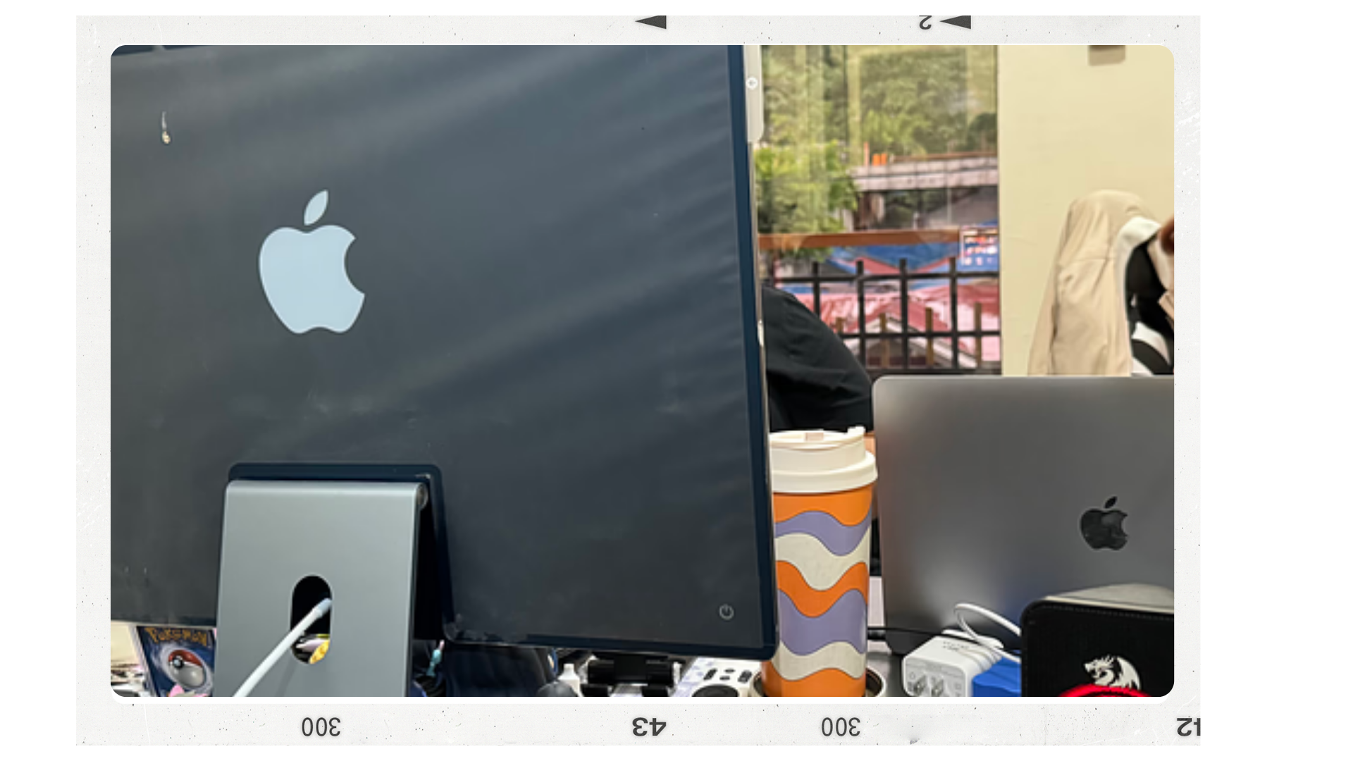 Two Apple computers on a cluttered desk, one facing away with the Apple logo visible, and a colorful cup with a wavy pattern between them, with background seen through a window.