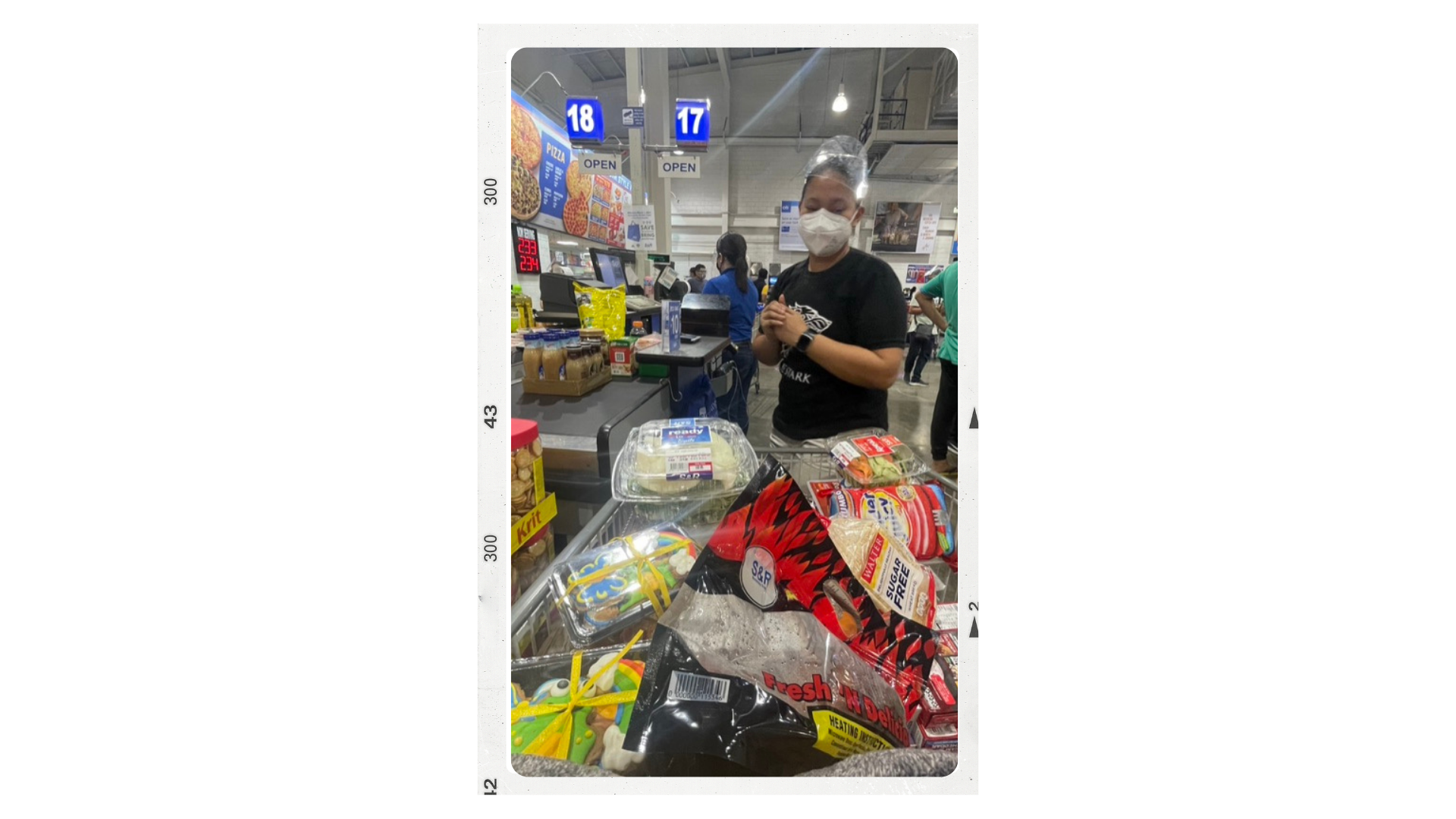Customer at the checkout counter in a grocery store, wearing a face mask and black T-shirt, with grocery items in the cart including packaged baked goods, candies, and vegetables.