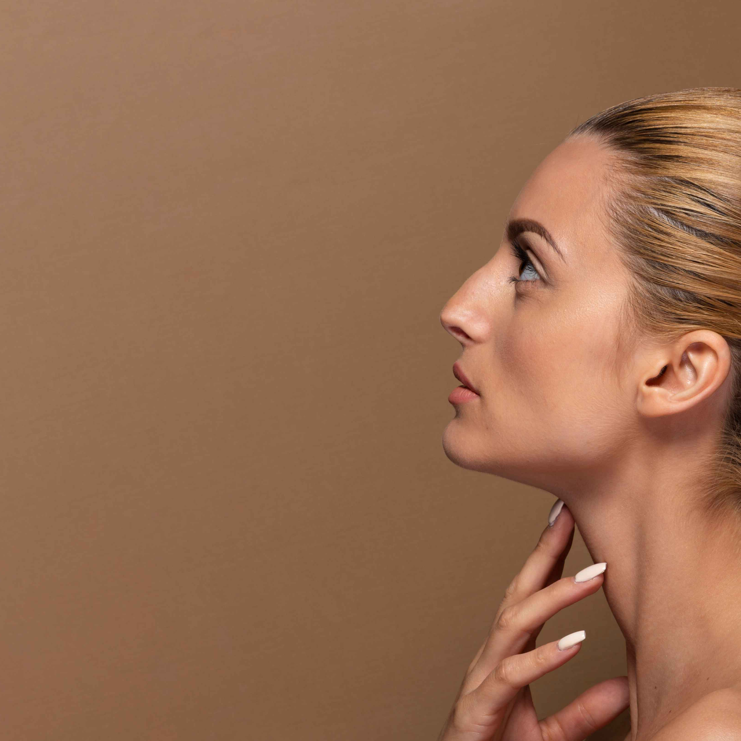 Side profile of a woman with blonde hair and clear skin touching her neck with her right hand, against a neutral brown background.