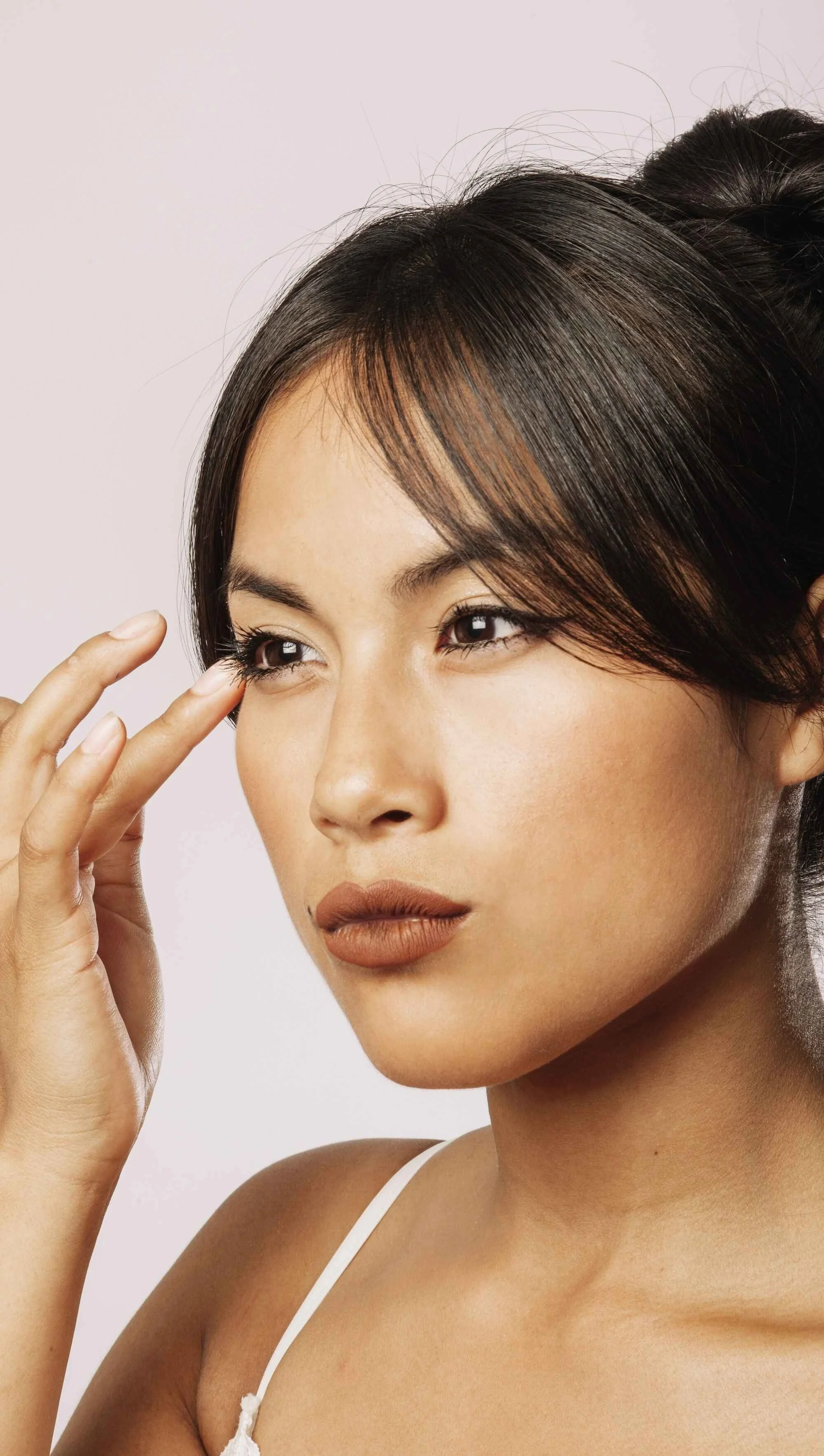 Close-up of a woman with styled dark hair, wearing makeup, and touching her temple with her hand, against a plain light background.