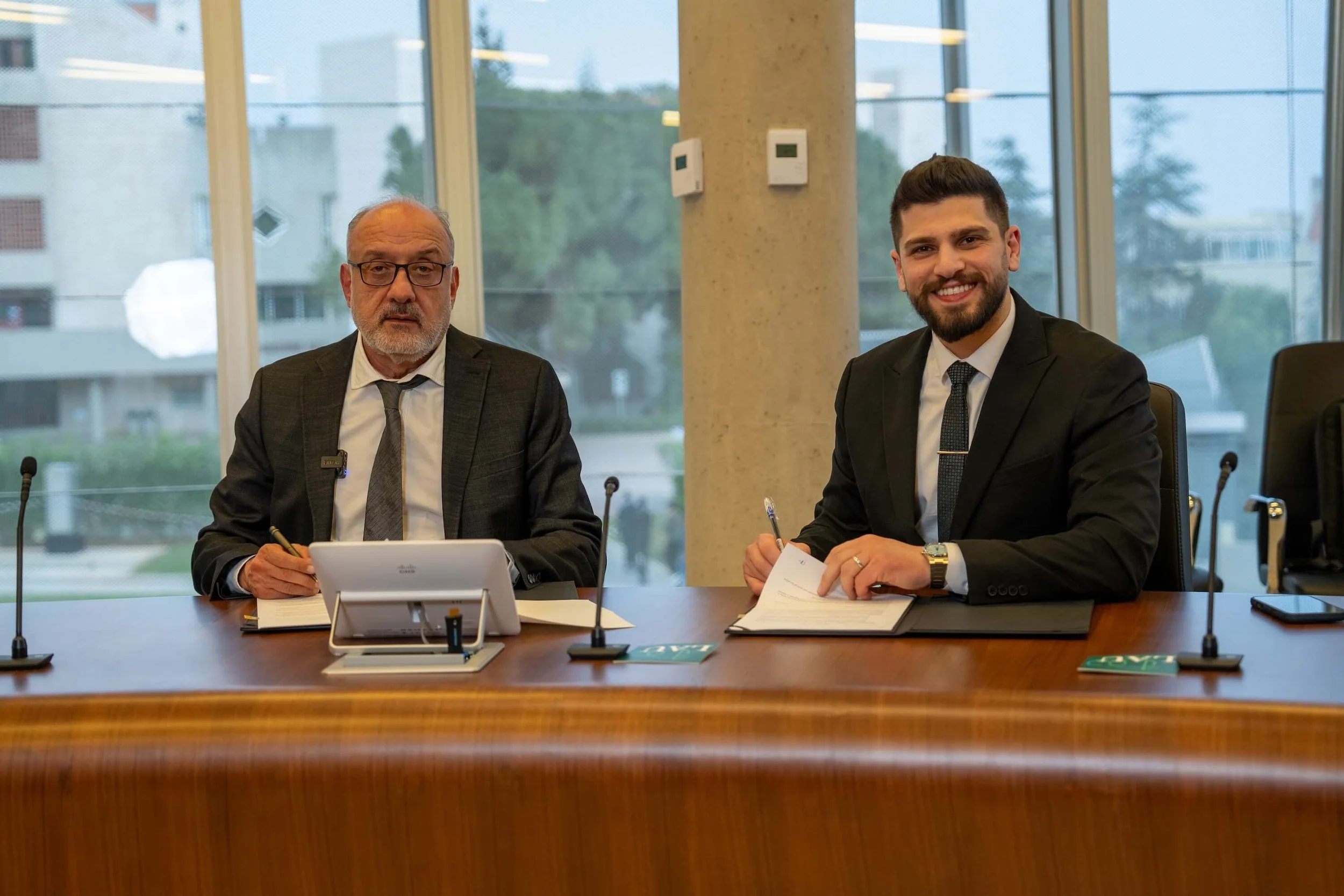 Two men in business suits sitting at a conference table. The older man on the left is wearing glasses, a dark suit, and a patterned tie, with a notepad and pen. The younger man on the right has a beard, a dark suit, a white shirt, and a tie, with a notepad and pen, smiling at the camera. The room has large windows in the background.