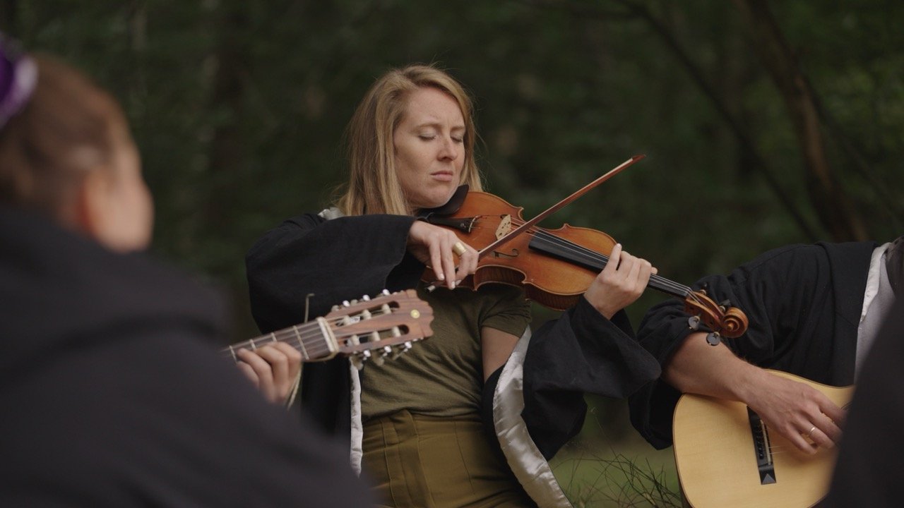 A woman with reddish hair playing a violin outdoors, surrounded by blurred figures holding guitars, in a forest setting.