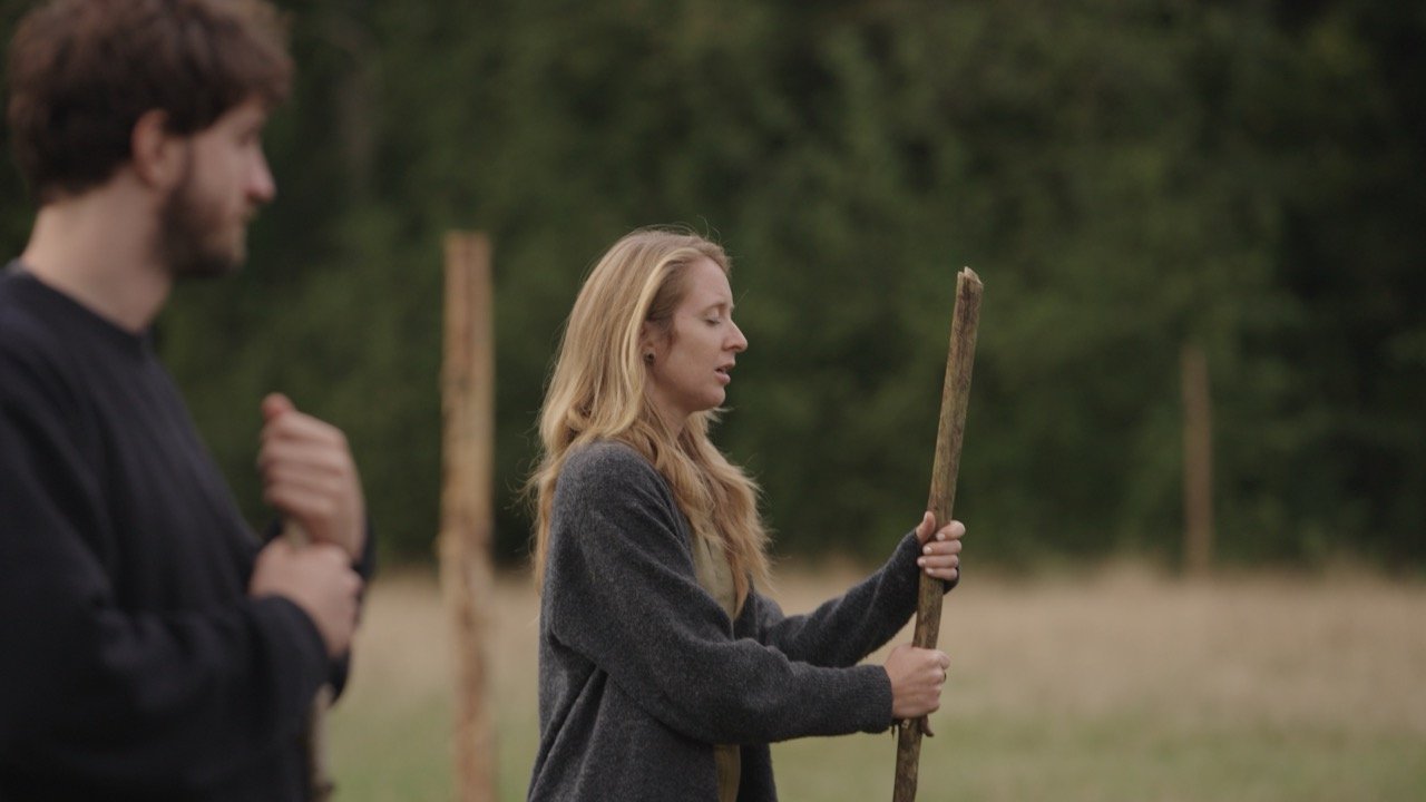 A blonde woman holding a stick standing outdoors in a field with trees in the background. A man with dark hair and beard is blurred in the foreground, appearing to hold or adjust his shirt.