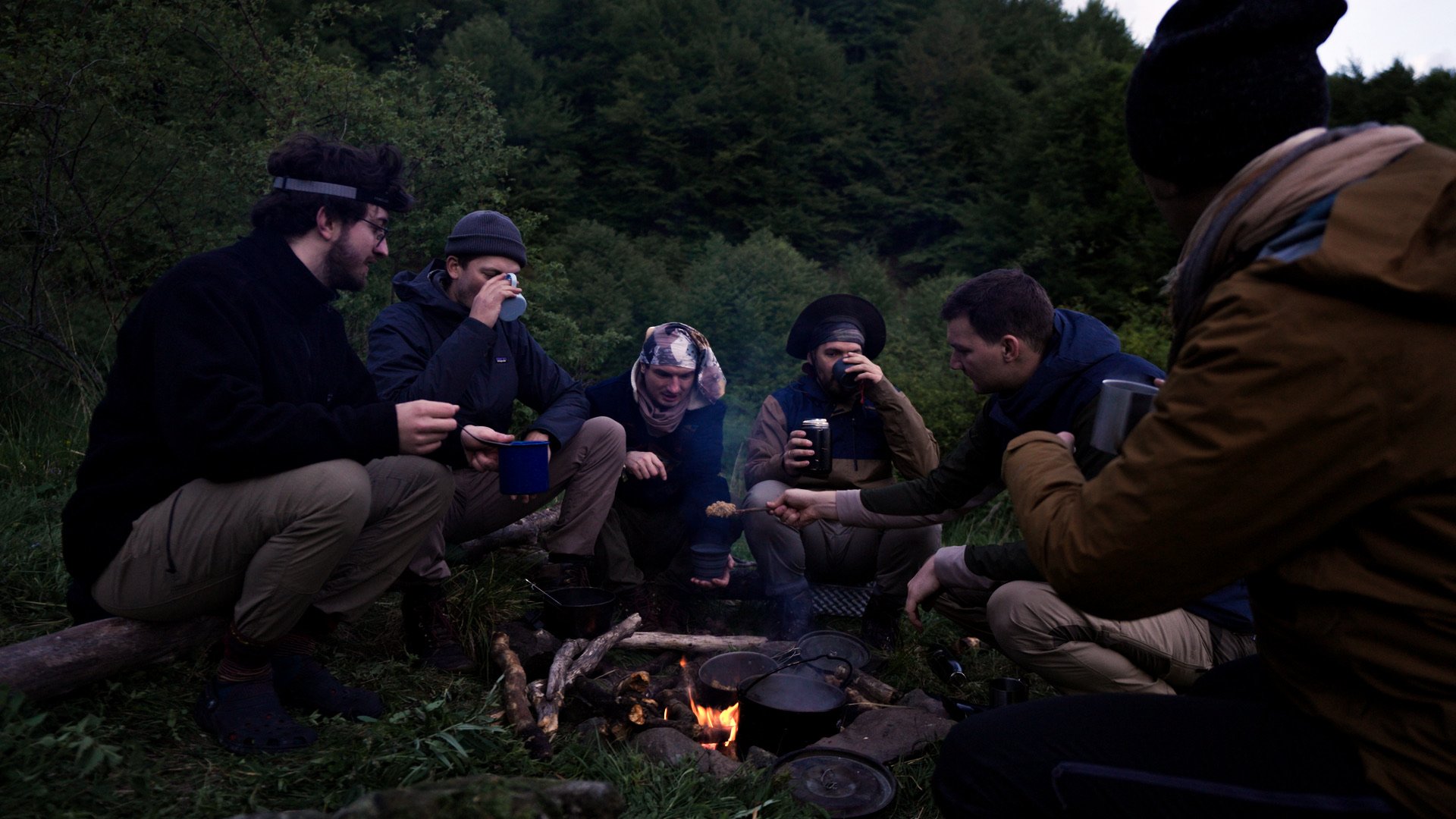 Six people sitting around a campfire in a forest, with camping cookware and drinks, enjoying the evening outdoors.