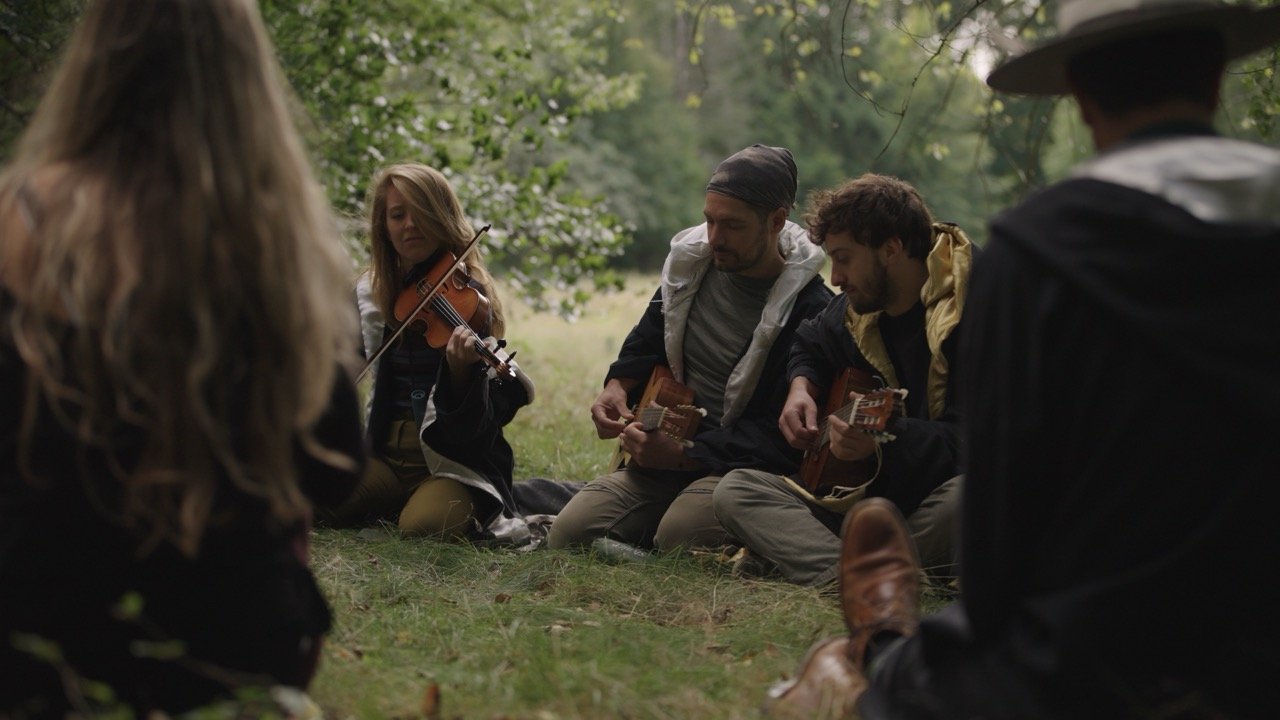 People sitting on the grass in a wooded area, playing guitar and violin, enjoying an outdoor music session.