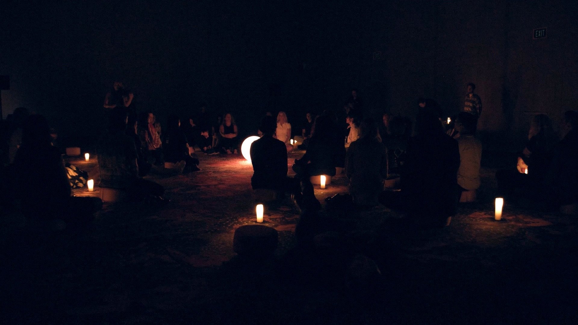People sitting on the floor in a dark room illuminated by candles and a glowing moon-shaped light, participating in a group meditation or gathering.
