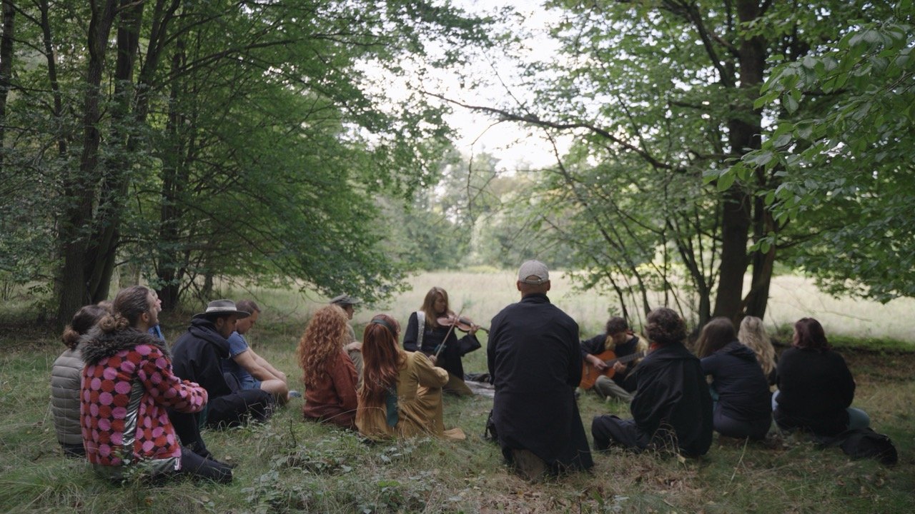 People sitting cross-legged and facing a woman playing a violin in an outdoor setting with trees and grass.