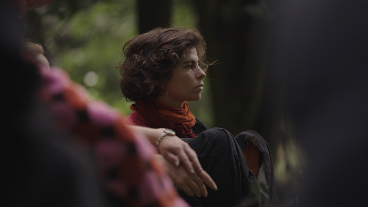 A woman with short curly hair and a red scarf around her neck sitting outdoors in a forested area, gazing thoughtfully to the right.