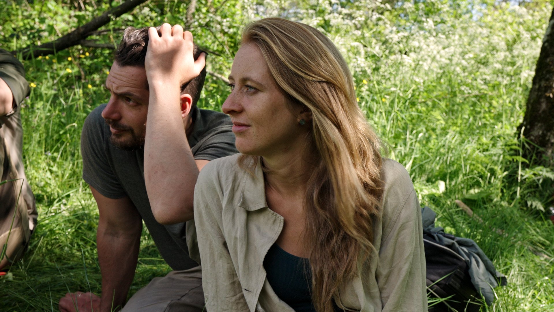 A woman with long, blonde hair sitting outdoors in a grassy area, gazing to the right with a thoughtful expression, while a man with dark hair and beard, wearing a gray shirt, is lying beside her with his hand on his head, looking upset, in a lush green environment.