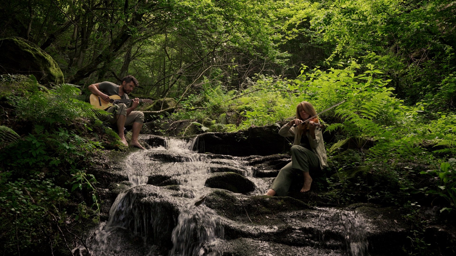 A man playing guitar and a woman playing violin sitting on rocks by a small waterfall in a lush green forest.