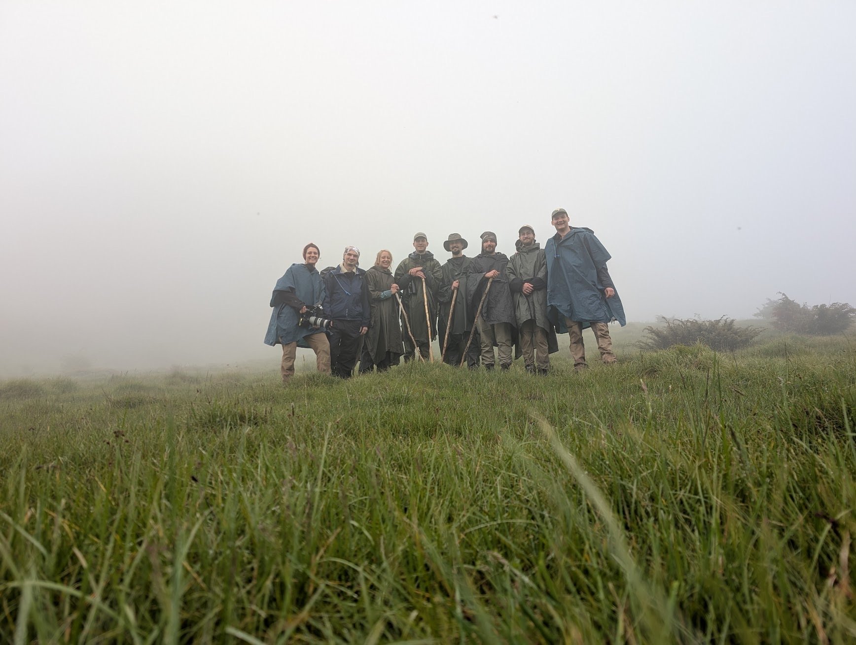 Group of nine people standing together on a grassy field in foggy weather, dressed in rain ponchos and casual outdoor clothing, some holding walking sticks, with some holding cameras.