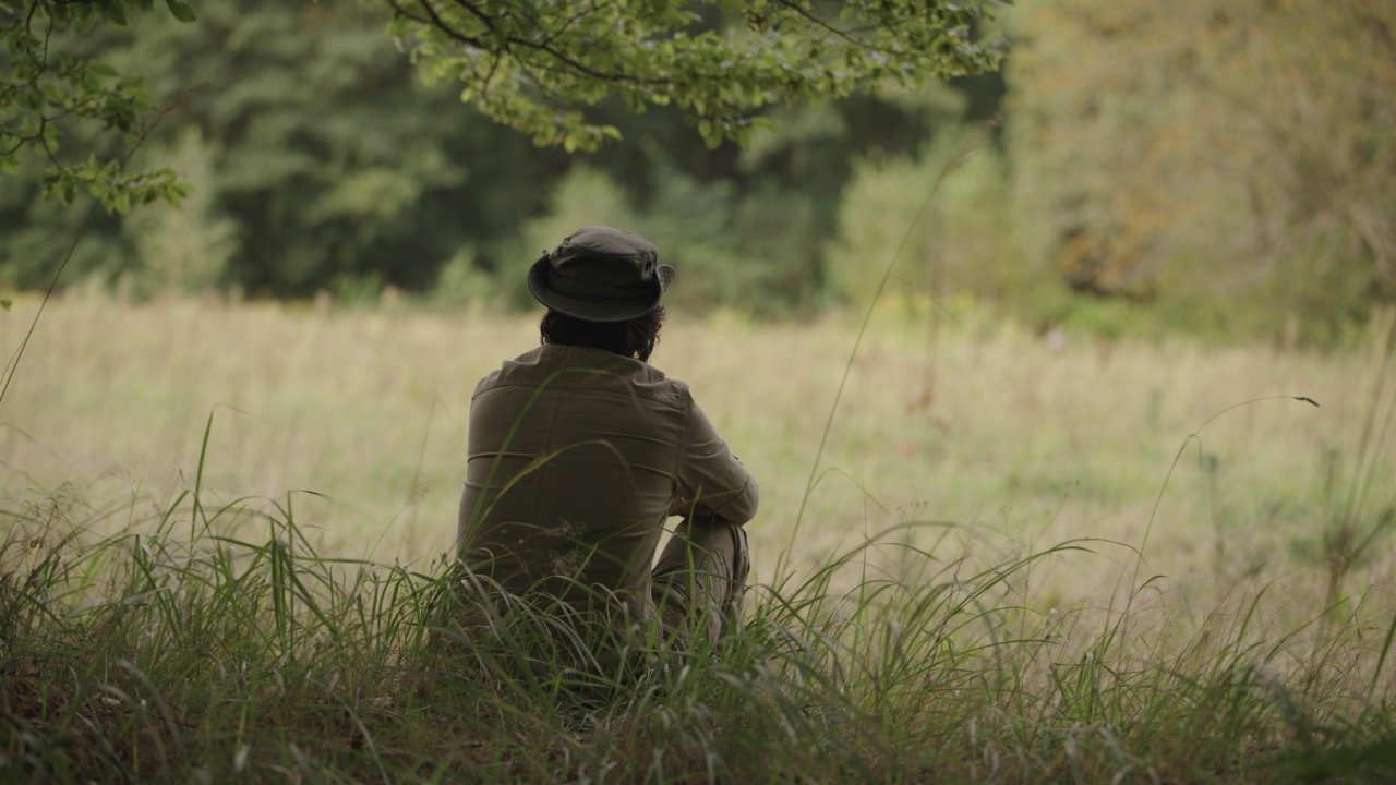A person sitting on the grass under a tree, looking out into a field, wearing a hat and outdoor clothing.
