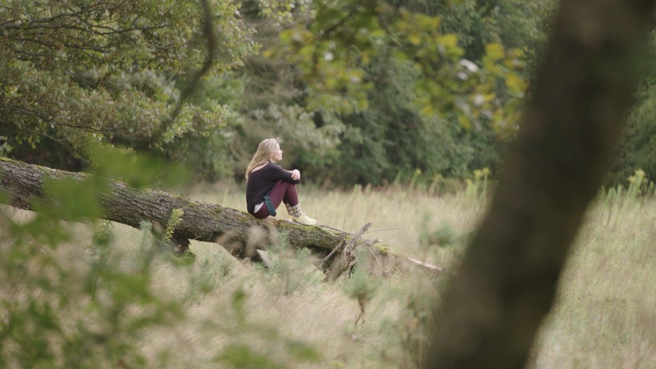 A girl sitting on a fallen tree trunk in a grassy, wooded area.