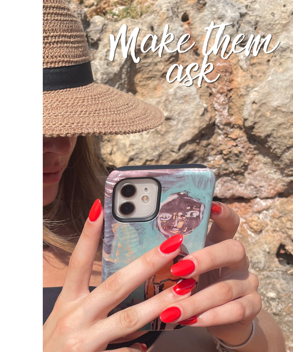 A woman wearing a brown textured wide-brim hat with a black band, and a black top, taking a selfie with her phone in front of a rocky background. She has painted bright red nails and is looking at her phone. There is a statement in white text that says 'Make them ask'.