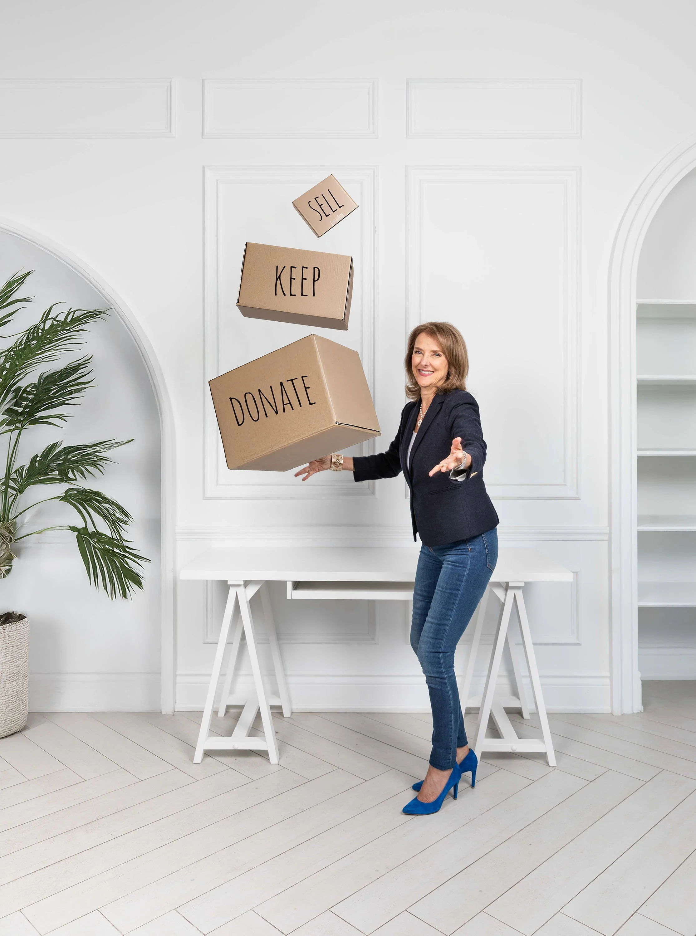 A woman in a black blazer, jeans, and blue high heels is tossing cardboard boxes with the words 'SELL,' 'KEEP,' and 'DONATE' written on them into the air inside a bright white room with decorative wall paneling and built-in shelves.