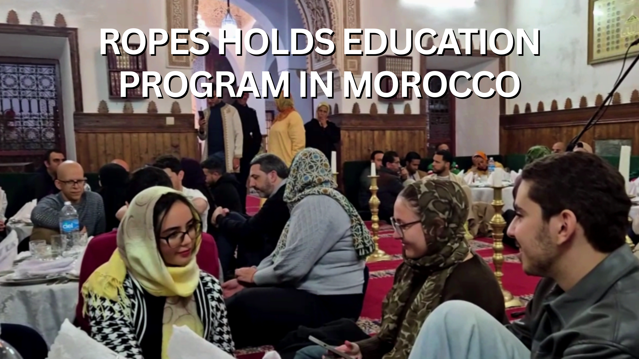 A diverse group of people attending a cultural educational event in Morocco, sitting on the floor and around tables, with traditional Moroccan decor and architecture visible in the background, including wooden paneling, arches, and ornate candle holders.