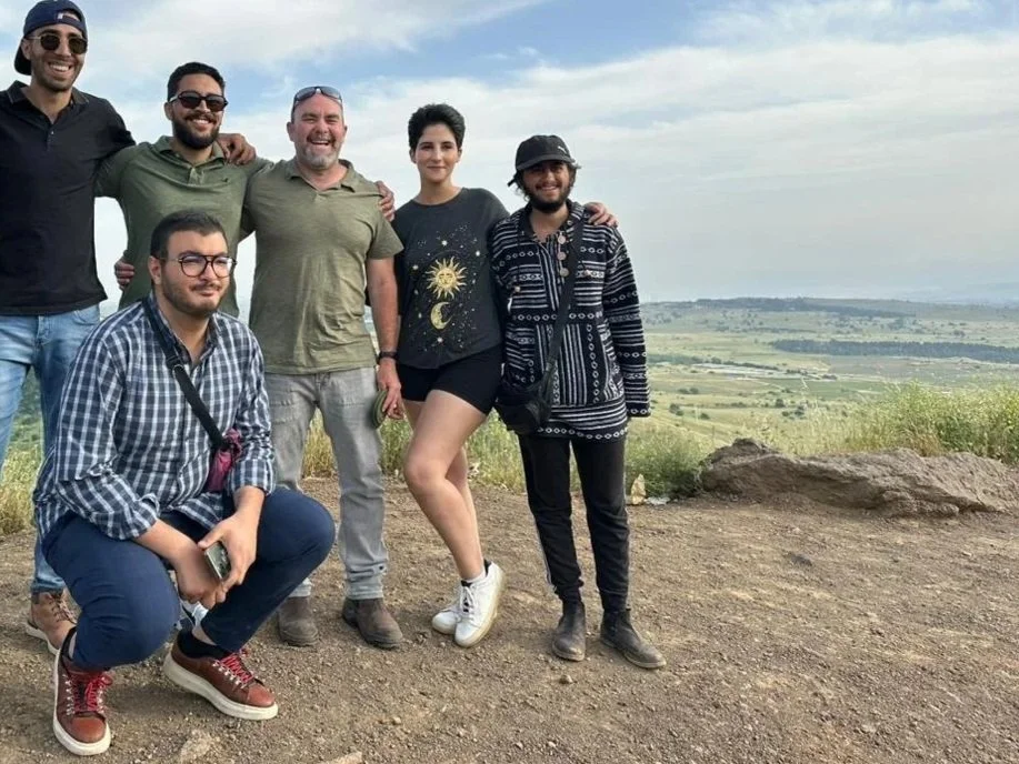 Group of eight friends posing outdoors on a hilltop with a scenic landscape of rolling hills and cloudy sky in the background.