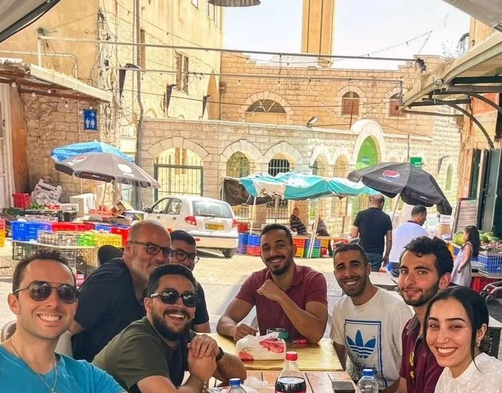 Group of nine people sitting at a wooden table outdoors, smiling at the camera, with a street scene and buildings in the background.