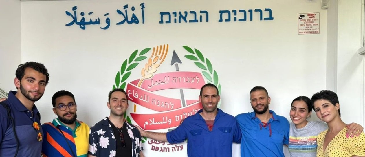 A group of nine people standing in front of a wall with Hebrew, Arabic, and English text. They are smiling and posing for the photo, with some individuals wearing casual and colorful clothing. The wall behind them has a graphic design with a laurel w