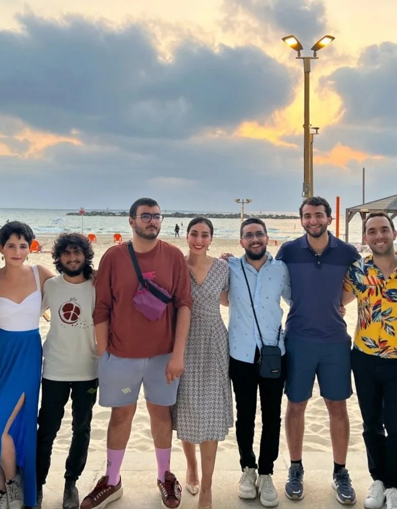 Group of nine friends smiling and posing on a beach at sunset, with sand, ocean, and cloudy sky in the background.