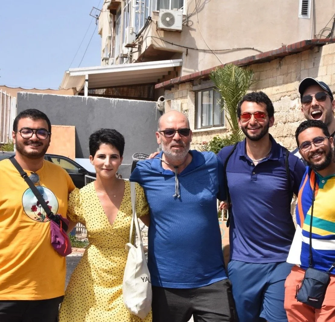 Group of eight diverse friends smiling and posing for a photo outdoors on a sunny day with a beige building, a palm tree, and street signs in the background.