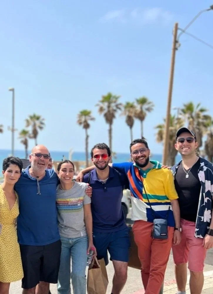 Group of ten people smiling outdoors near a beach with palm trees, parked cars, and a blue sky in the background.