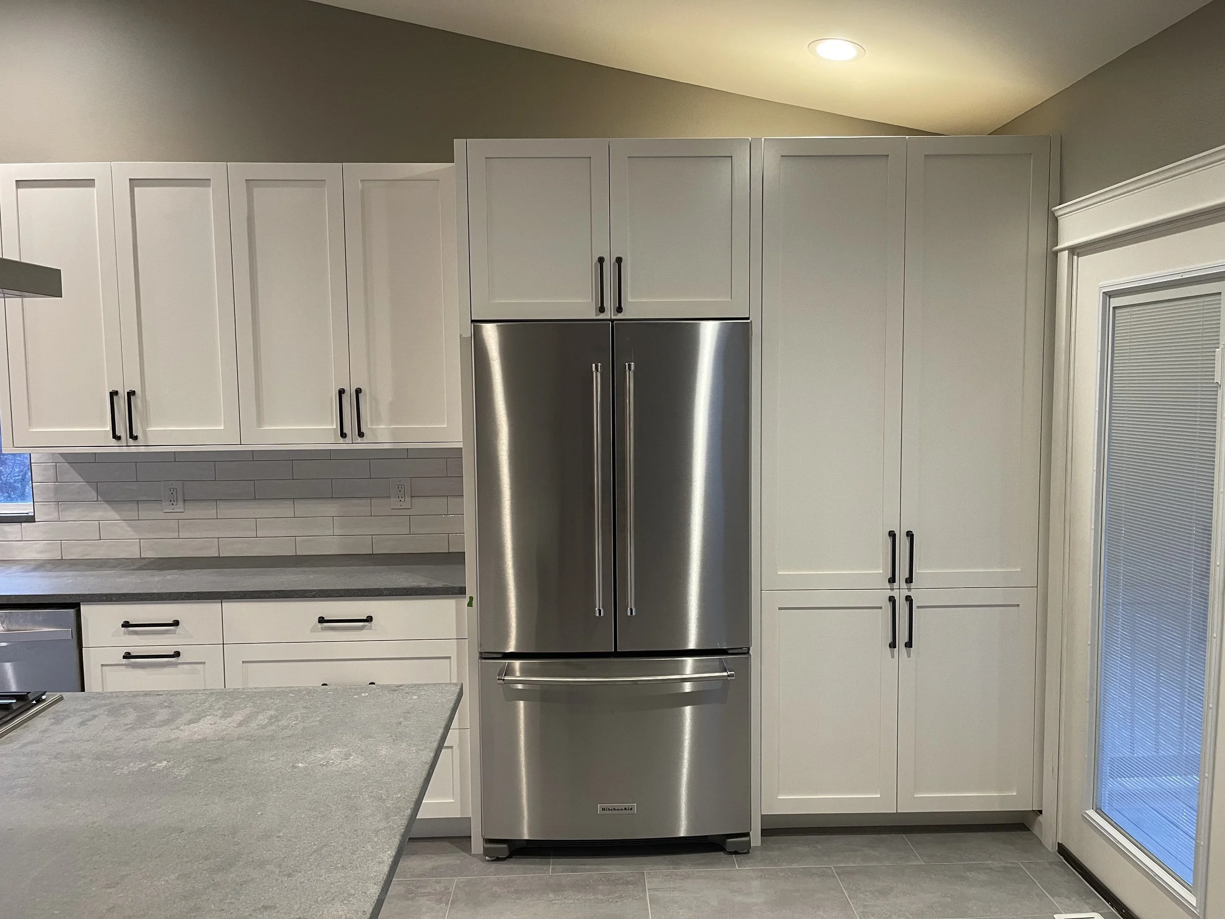 Tall pantry cabinets and fridge surround in a white shaker kitchen with matte black pulls—Noah & Noble custom cabinetry.