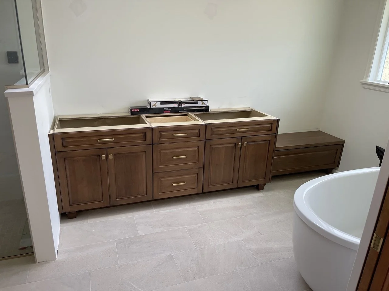 In-progress primary bath: custom double-sink vanity in warm stained wood with raised-panel doors, center drawer stack, brass hardware, and matching low bench—Noah & Noble, Greater Seattle.