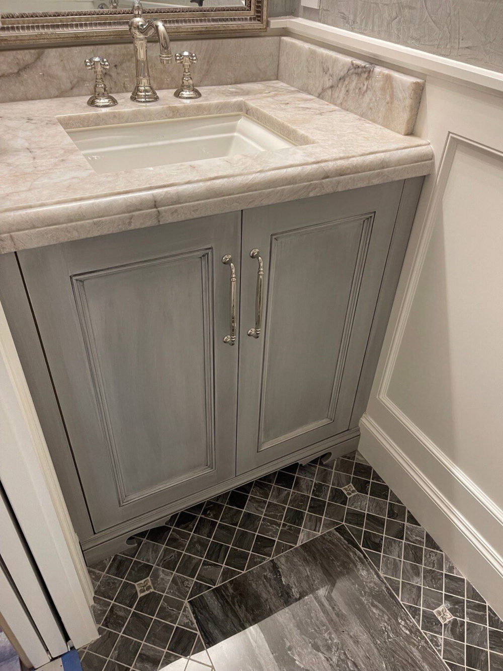 Custom powder room vanity with gray hand-applied finish, framed-panel doors, polished-nickel pulls, and marble top with backsplash—Noah & Noble.