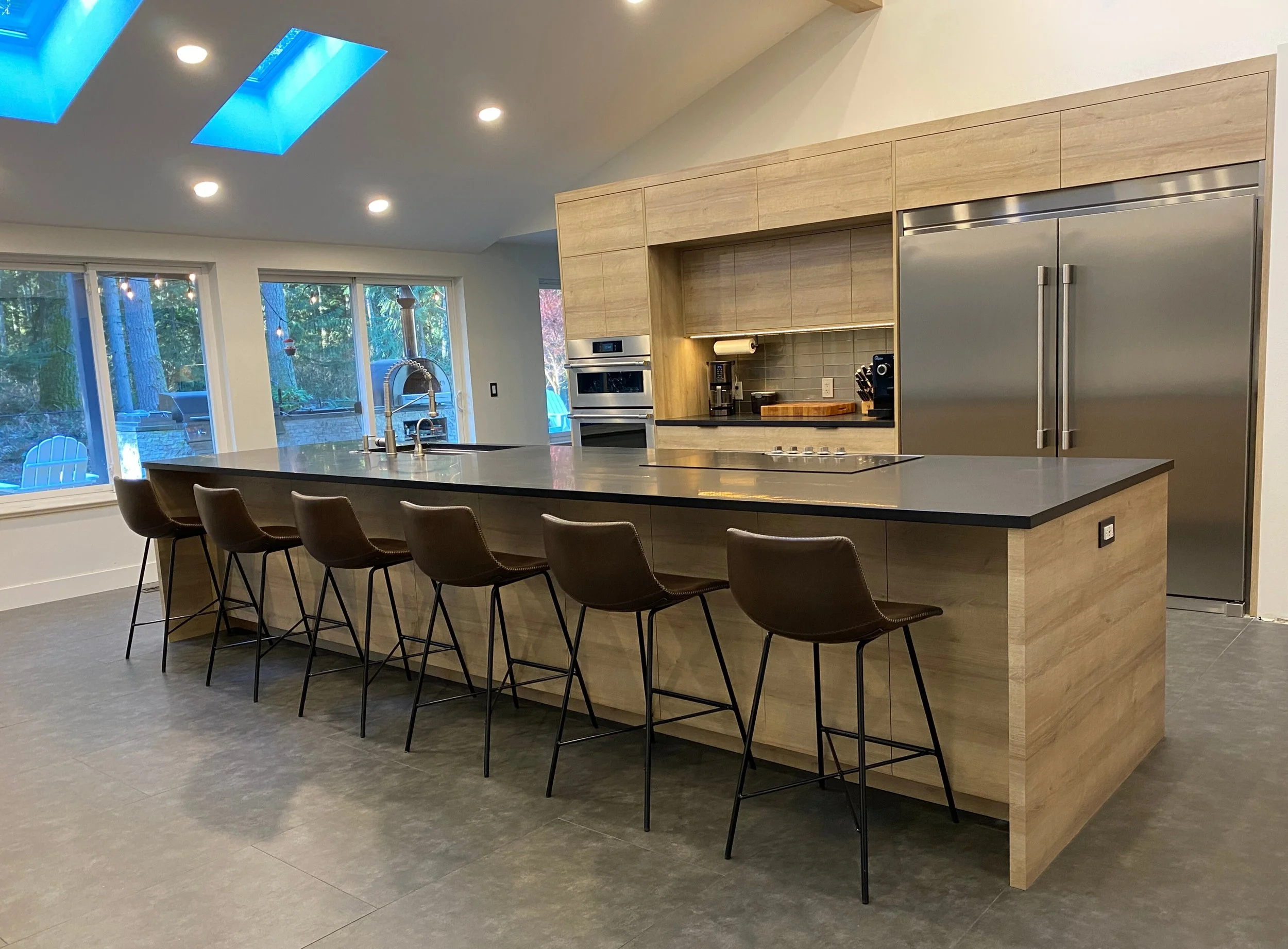Oversized waterfall-free island with dark quartz top, bar seating for seven, prep sink and cooktop, facing a full-height appliance wall—Noah & Noble custom kitchen.