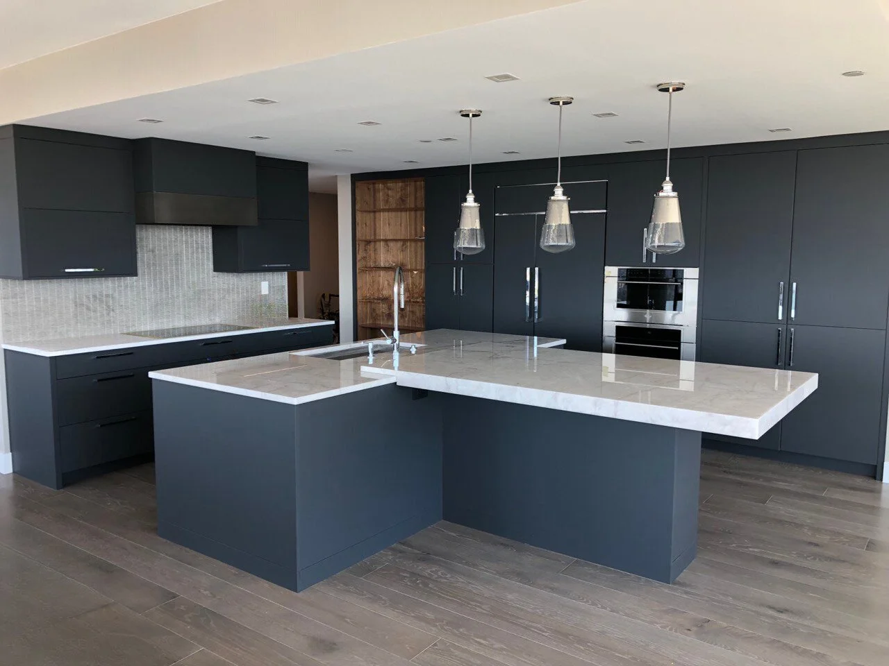 Modern charcoal kitchen with slab-front cabinets, full-height pantry wall, panel-ready fridge, wall ovens, and an oversized island with thick mitered-edge stone top—Noah & Noble, Greater Seattle.