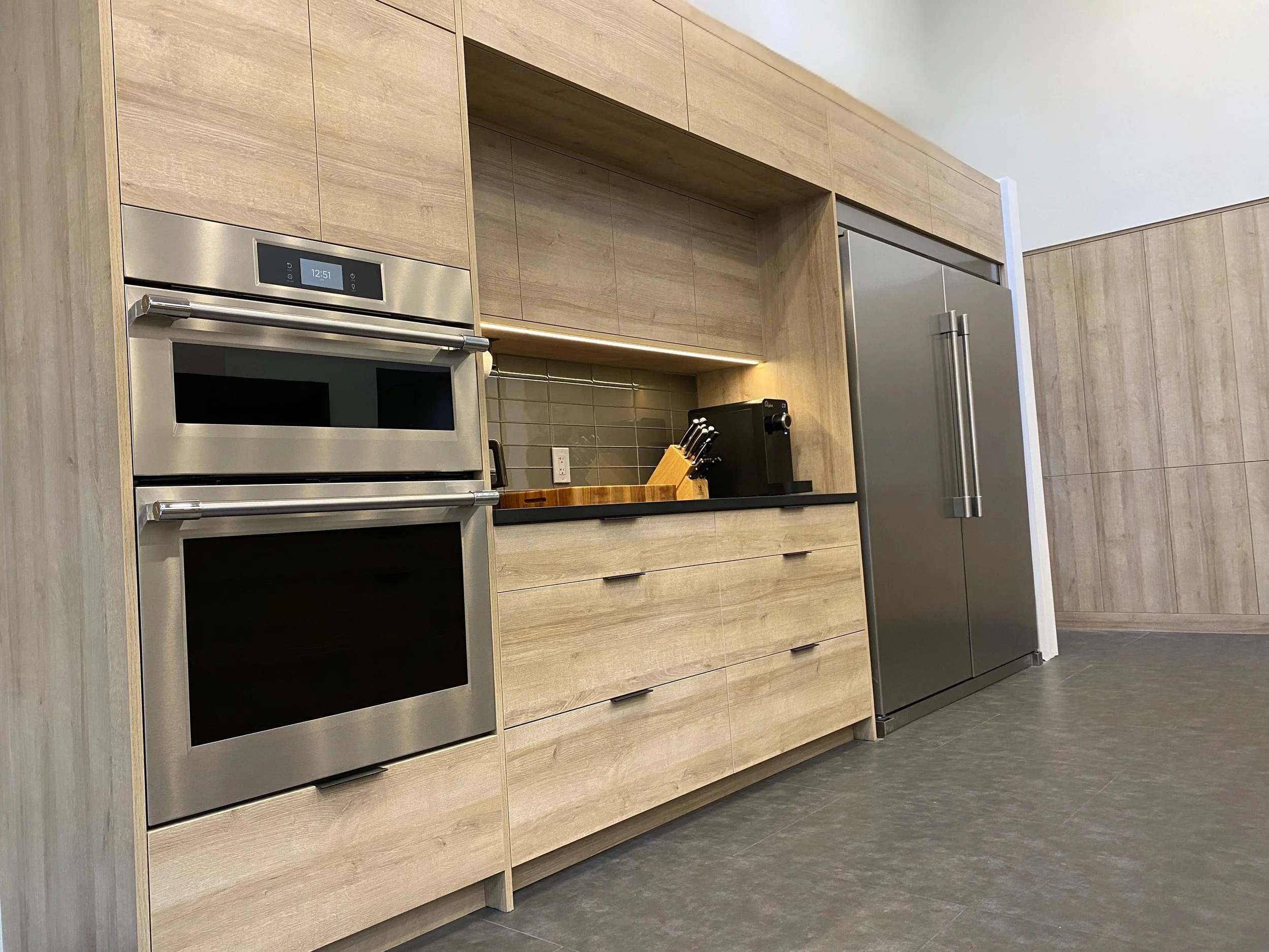 Modern kitchen wall with light wood slab cabinets, double wall ovens, appliance niche with under-cabinet lighting, and built-in fridge—Noah & Noble.