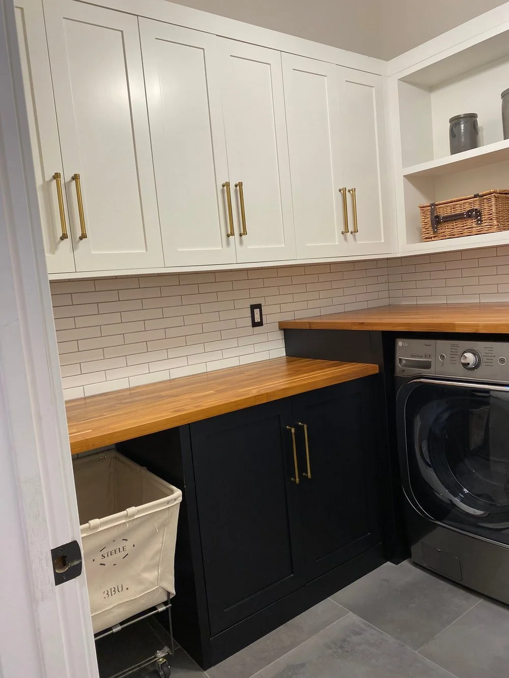 Two-tone laundry room cabinets—white shaker uppers with brass pulls, black base cabinets, butcher-block counter, subway-tile backsplash, open shelf, and front-load washer—Noah & Noble, Greater Seattle.