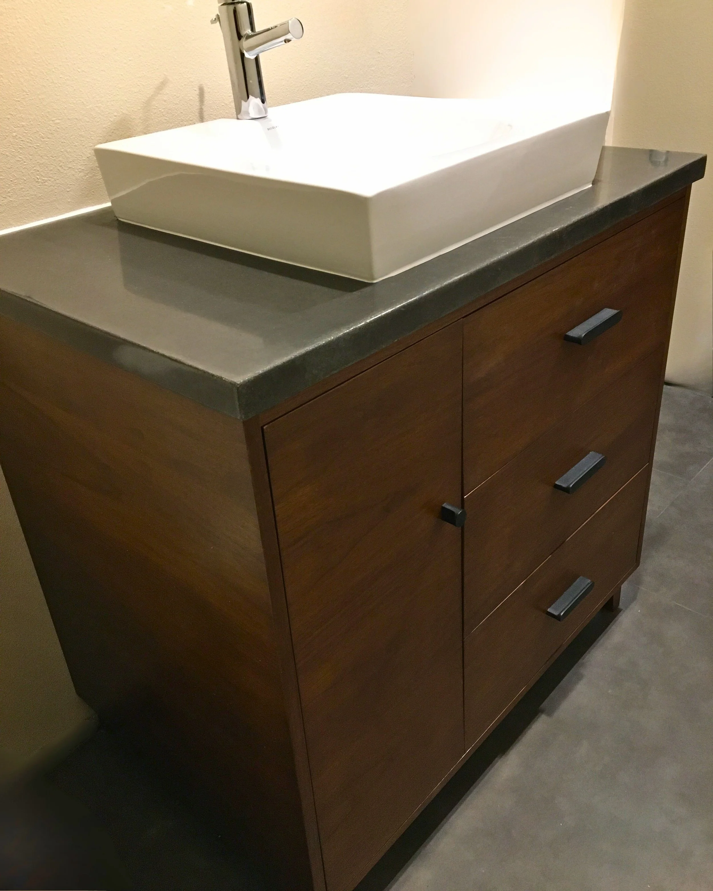 Modern walnut bathroom vanity with slab-front drawers, matte black pulls, polished concrete countertop, and white vessel sink—Noah & Noble, Greater Seattle.