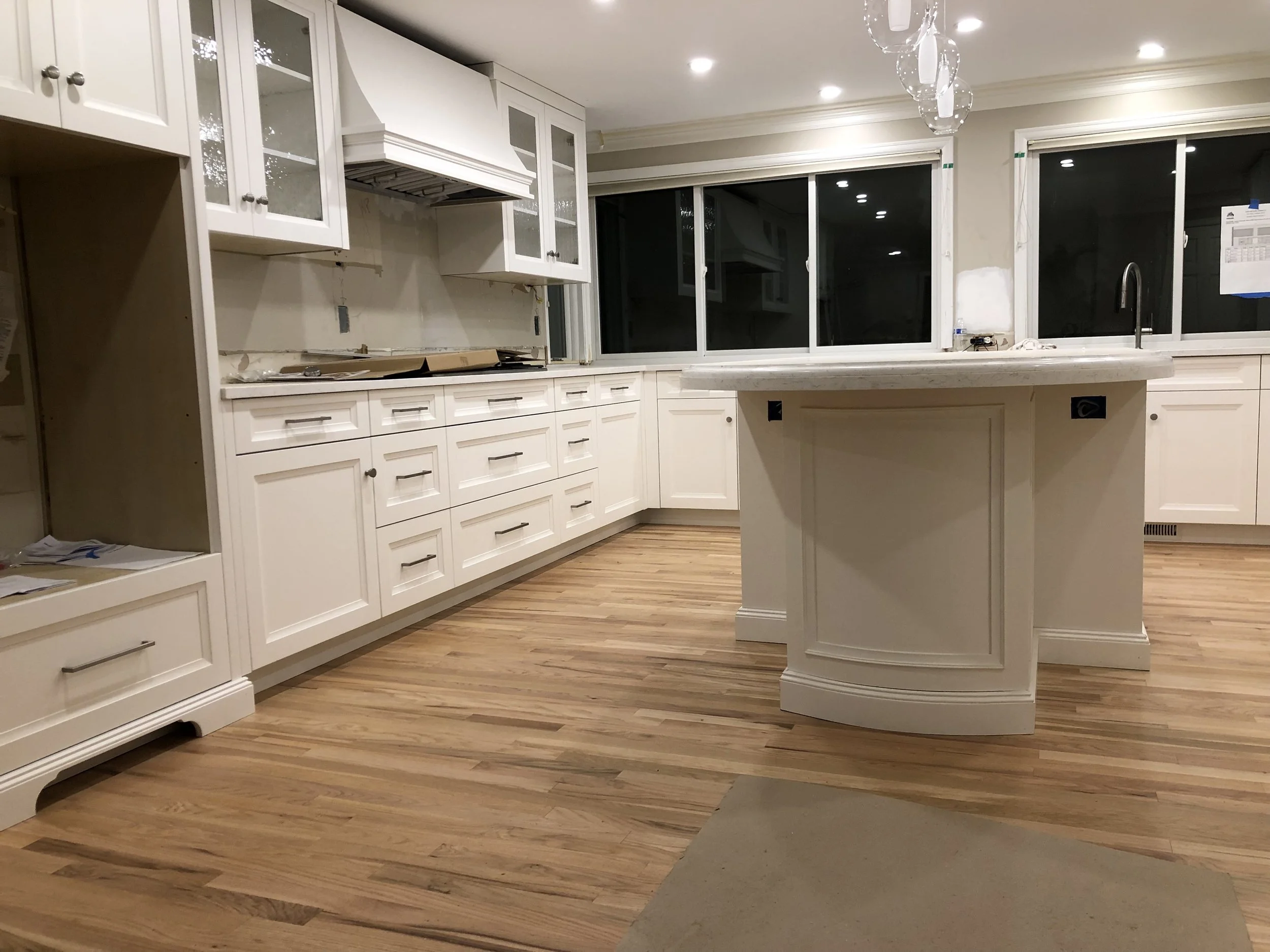 Wide view of custom white shaker kitchen with long drawer run, glass-front uppers, integrated range hood, and a curved island with stone top—Noah & Noble, Greater Seattle.