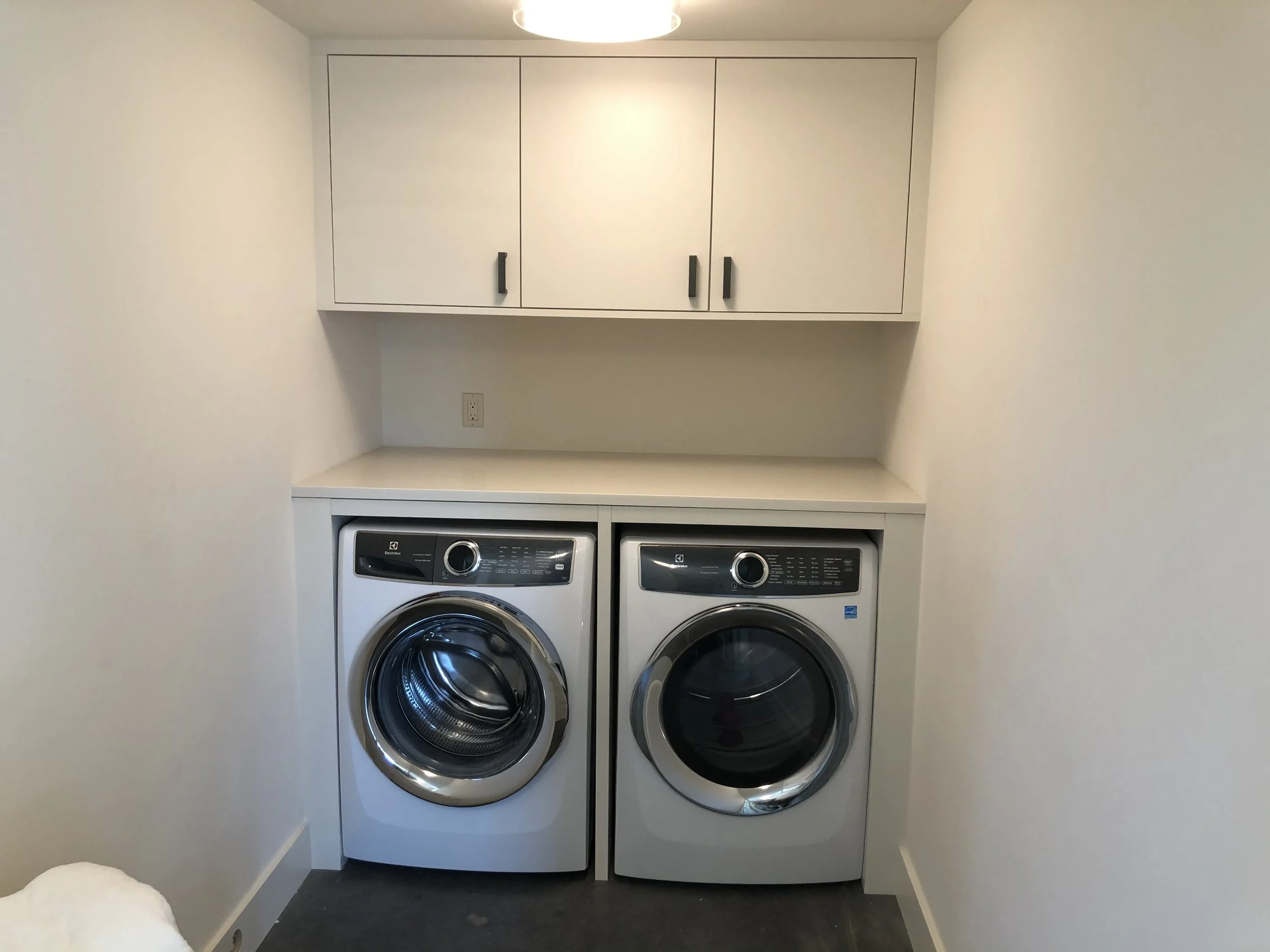 Modern laundry wall: white upper cabinets, open shelf, and full-width quartz countertop spanning front-load machines—custom cabinetry by Noah & Noble.