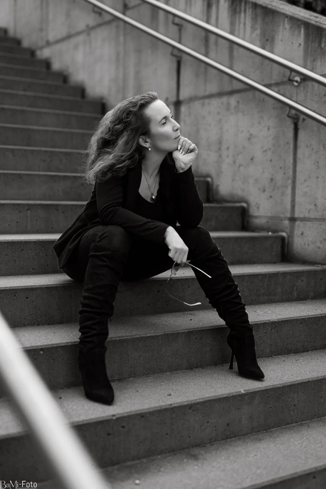 A woman sitting on urban concrete stairs, resting her chin on her hand, holding sunglasses in her other hand, in a thoughtful pose, depicted in black and white.