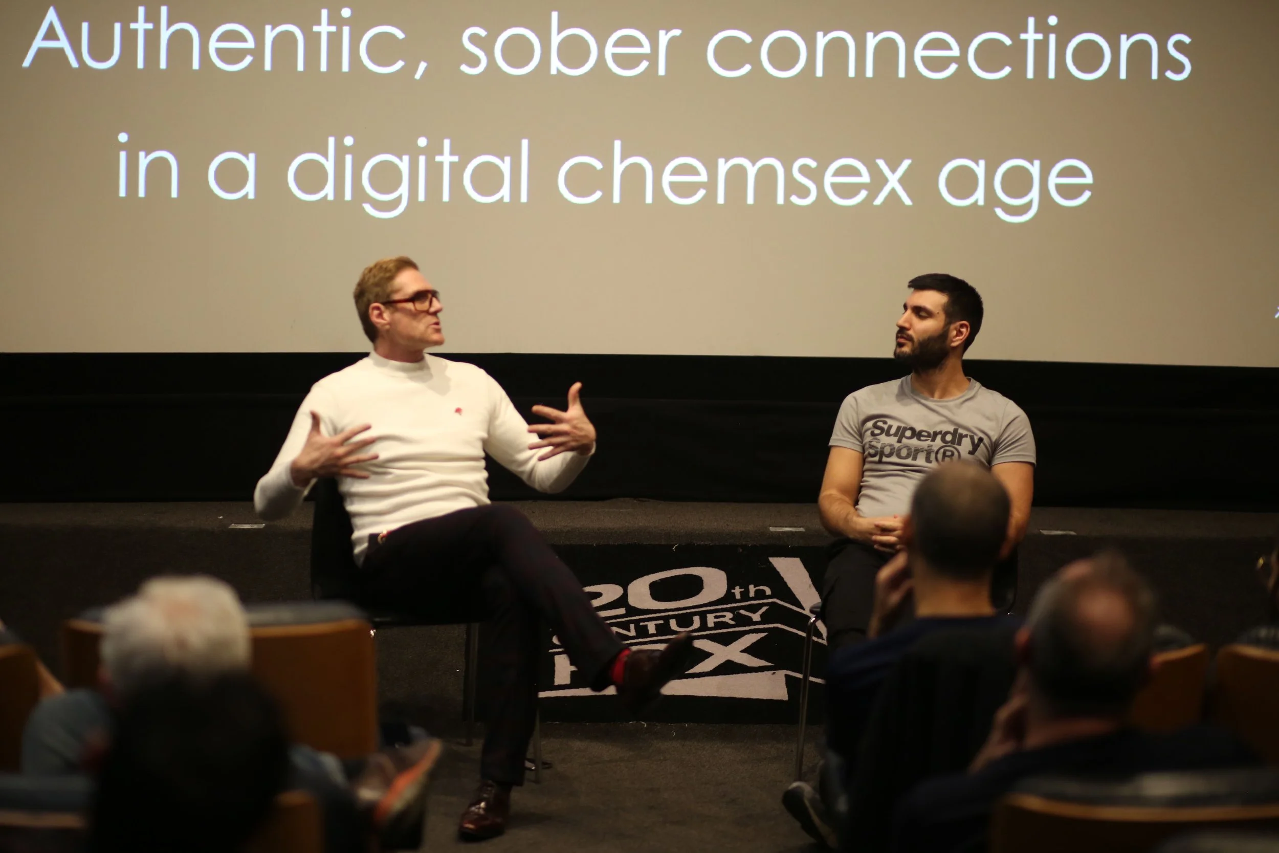 Two men are sitting on stage in front of an audience, engaged in a discussion. A large screen behind them displays the text 'Authentic, sober connections in a digital chemsex age.' The man on the left wears glasses and a white shirt, gesturing as he speaks. The man on the right wears a gray Superdry Sport T-shirt, listening attentively. Audience members are visible in the foreground.