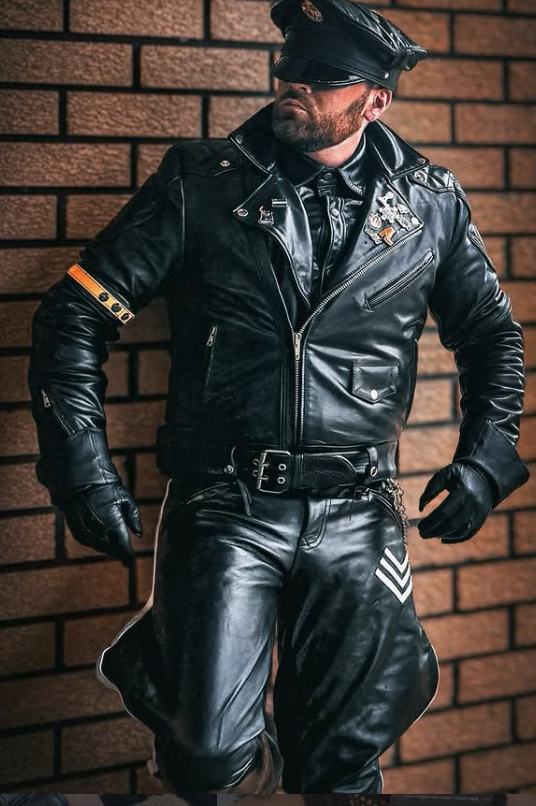 A man in a black leather outfit, including a leather jacket, leather pants, and leather gloves, stands against a brick wall. He wears a black cap and sunglasses, giving a confident pose.